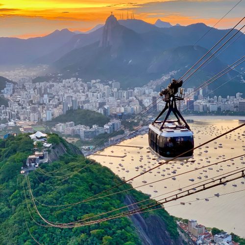 Sugarloaf mountain cable car during sunset in Rio de Janeiro, with Christ the Redeemer statue and boats in the background.